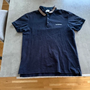 Svart pikétröja från Burberry - Snygg svart pikétröja från Burberry med klassisk krage och knappar. Tröjan har ett broderat Burberry-märke på bröstet och randiga detaljer på kragen i beige, röd och svart. Perfekt för en stilren look.  Nypris 4990kr