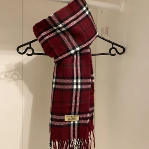 Burberry - Snygg rutig halsduk från Burberry i en djup burgendy röd färg. Tillverkad i 100% lammull med klassiskt fransad kant. Den är i mycket fint skick utan några defekter. Perfekt för att hålla dig varm och stilren under kyliga dagar.