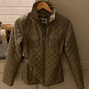 Olivgrön quiltad ridjacka från CRW - Snygg ljusgrön ridjacka som passar bra till våren.  Jackan är i mycket bra skick och ser nästan ny ut.  Jackan är i storlek XXS för barn från 10-13 år.  Man kan justera storleken i midjan och man kan öppna nerifrån och uppifrån på dragkedjan så den passar när man sitter i sadeln. 