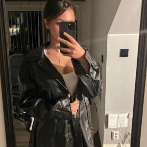 Svart skinnjacka med knytbälte - Snygg svart skinnjacka med knytbälte och långa ärmar. Perfekt för en stilren look. Jackan har en klassisk design med knappar framtill.