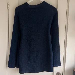 Blå glittrig klänning - Snygg blå glittrig klänning med öppen rygg och knytning i nacken. Perfekt för en festlig kväll. Klänningen har långa ärmar och en elegant passform.