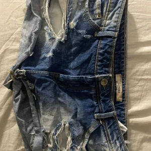 Blå jeansshorts från Bershka - Säljer ett par blå jeansshorts från Bershka med slitna detaljer och fransar. Shortsen har en klassisk femficksdesign och är perfekta för en avslappnad stil.