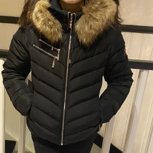 Svart dunjacka från Hollies - Snygg svart dunjacka från Hollies med pälskantad huva och dragkedja. Jackan har ett leopardmönstrat foder och flera praktiska fickor med dragkedjor. 