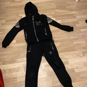 Svarta mjukisbyxor från Philipp Plein - Svarta mjukisbyxor med dragkedjor och snörning i midjan. Byxorna har en cool design med ett märke på benet och matchar perfekt med en hoodie från samma märke.
