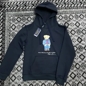 Svart hoodie från Polo Ralph Lauren - Snygg mörkblå  hoodie från Polo Ralph Lauren med en ikonisk björn på framsidan. Perfekt för en avslappnad stil med en touch av lyx. Den har en klassisk känguruficka och justerbar huva.