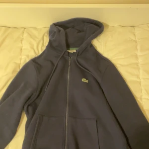 Mörkblå hoodie från Lacoste - Säljer en mörkblå hoodie från Lacoste med dragkedja. Tröjan har en bekväm passform och är perfekt. Den är i storlek m men passar perfekt i s. Pris kan diskuteras.