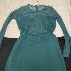 Grön klänning från VRS Woman - Elegant grön klänning med långärmad design och spetsdetaljer på överdelen. Klänningen har en dragkedja i ryggen och en rund halsringning med en liten öppning. Perfekt för en stilfull look.