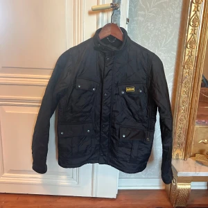 Svart quiltad jacka från Barbour storlek XL till barn - Snygg svart quiltad jacka från Barbour International med dragkedja och knappar. Jackan har flera praktiska fickor och en stilren design. Jackan är i mycket bra skick! Använd ett fåtal gånger. Storlek XL TILL BARN . Motsvarar ca 11-12 år.