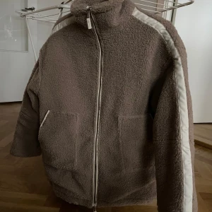 Beige teddyjacka med dragkedja - Mysig beige teddyjacka med dragkedja och vita detaljer längs ärmarna. Jackan har en hög krage och två framfickor, perfekt för kyliga dagar. Helt oanvänd 