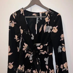 Blommig klänning med knytband - Elegant svart klänning med blommönster i rosa och grått. Klänningen har långa ärmar och ett knytband i midjan för en smickrande passform. Perfekt för både vardag och festliga tillfällen.