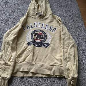 Säljer en superfin beige/ljusgul hoodie från Falsterbo Horse Show. Den har en stor logga på framsidan med texten 'Falsterbo Horse Show' och en häst i mitten. Hoodien är i storlek S och har en mysig huva. Perfekt för en avslappnad stil eller för att visa din kärlek till hästsport!