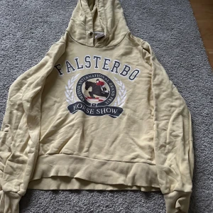 Beige hoodie från Falsterbo Horse Show - Säljer en superfin beige/ljusgul hoodie från Falsterbo Horse Show. Den har en stor logga på framsidan med texten 'Falsterbo Horse Show' och en häst i mitten. Hoodien är i storlek S och har en mysig huva. Perfekt för en avslappnad stil eller för att visa din kärlek till hästsport!