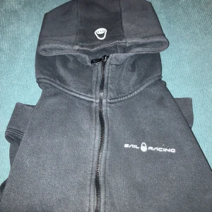 Svart hoodie från Sail Racing - Säljer en svart hoodie från Sail Racing i storlek 170. Den har en dragkedja framtill och en mysig huva. Perfekt för kyliga dagar och har praktiska fickor framtill. Passar både till vardags och för sportiga aktiviteter.