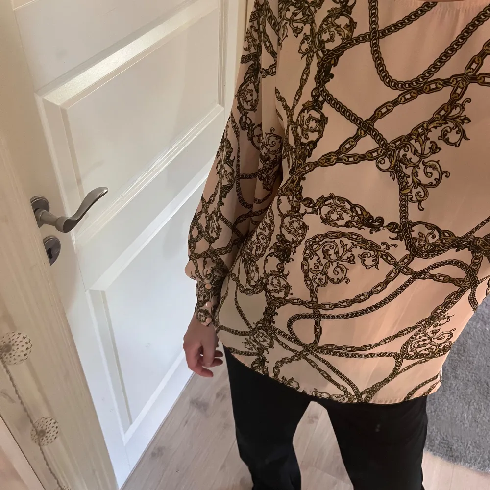 Säljer en snygg beige blus med ett coolt kedjemönster i svart. Blusen har långa ärmar och en lös passform, perfekt för både vardag och fest. Den är gjord i ett mjukt och bekvämt material som känns skönt mot huden.. Blusar.