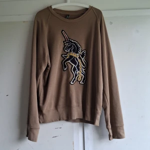 Beige tröja med enhörning från Neil Barrett - Säljer en snygg beige tröja från Neil Barrett med en enhörning på framsidan. Tröjan är långärmad och gjord av mjukt material, perfekt för höst och vinter. Den har en normal passform och är i nyskick. Perfekt för att ge din garderob en unik touch!