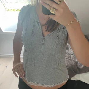 Grå ribbad topp med spetsdetaljer - Säljer en superfin grå ribbad topp med spetsdetaljer vid ärmslut och nederkant. Toppen har korta ärmar och en v-ringad knäppning framtill. Perfekt för en casual look eller att klä upp med smycken!