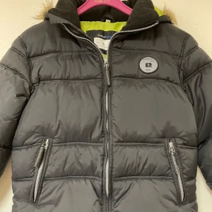 Svart pufferjacka med huva - Säljer en snygg svart pufferjacka. Jackan har en mysig huva med fuskpäls och dragkedja framtill. Perfekt för kalla vinterdagar! Den är i storlek 150 och har praktiska fickor med dragkedjor. Dock lite sliten då den har används några gånger, men fortfarande i fint skick! ❤️