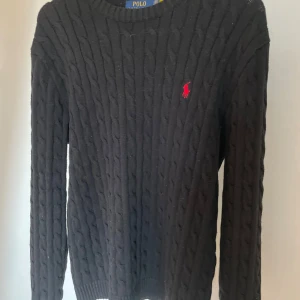 Svart kabelstickad tröja från Ralph Lauren - Säljer en stilren svart kabelstickad tröja från Ralph Lauren med det klassiska röda logotypen på bröstet. Tröjan har långa ärmar och en rund halsringning. Perfekt för kyligare dagar.