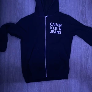 Svart hoodie från Calvin Klein Jeans - Säljer en stilren svart hoodie från Calvin Klein Jeans med dragkedja framtill. Tröjan har en klassisk design med huva och långa ärmar. Perfekt för en avslappnad look.