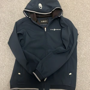 Mörkblå jacka från Sail Racing - Snygg mörkblå jacka från Sail Racing med dragkedja och huva. Jackan har långa ärmar och vita detaljer vid dragkedjan och ärmsluten. Perfekt för en stilren look.