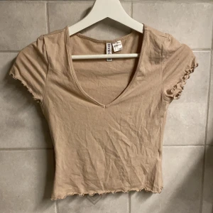 Beige v-ringad topp från H&M Divided - Säljer en beige v-ringad topp från H&M Divided. Toppen har korta ärmar och en fin volangdetalj vid ärmslut och nederkant. Perfekt för en avslappnad stil.