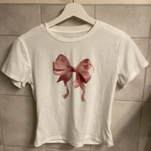 Vit t-shirt med rosa rosett - Säljer en söt vit t-shirt med en stor rosa rosett på framsidan. T-shirten har korta ärmar och en rund halsringning. Perfekt för en avslappnad och stilren look.