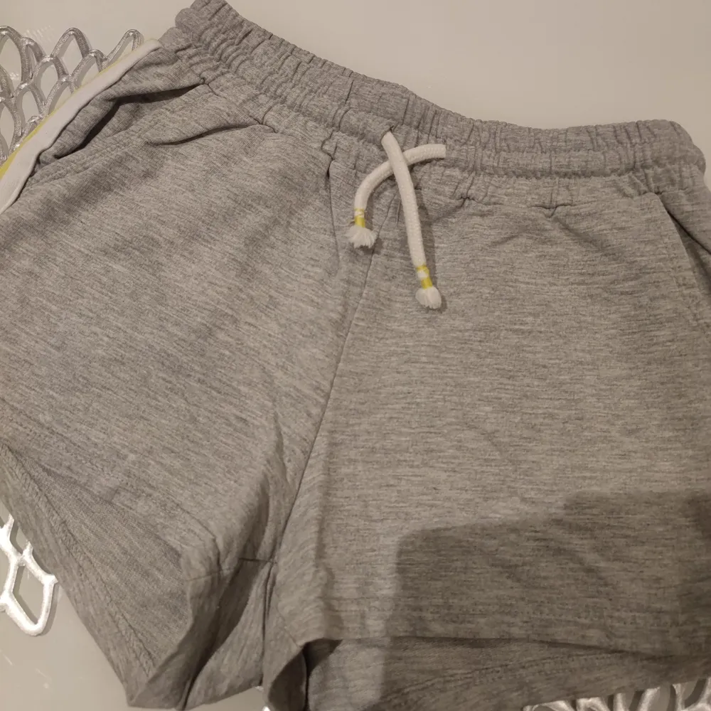 Snygga grå shorts från Lindex med elastisk midja och snörning. De har en vit och gul rand på sidan som ger en sportig look. Perfekta för en avslappnad stil.. Shortsit.