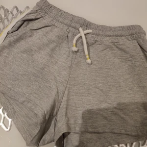 Grå shorts från Lindex - Snygga grå shorts från Lindex med elastisk midja och snörning. De har en vit och gul rand på sidan som ger en sportig look. Perfekta för en avslappnad stil.