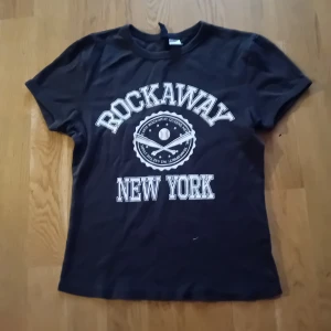 Svart t-shirt med Rockaway New York-tryck - Cool svart t-shirt med stort vitt tryck som säger 'Rockaway New York'. Perfekt för en avslappnad stil. Kortärmad och i mjukt material.