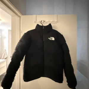 Svart teddyjacka från The North Face - Mysig svart teddyjacka från The North Face med dragkedja och hög krage. Perfekt för kyliga dagar med sin fluffiga och varma design. Jackan har långa ärmar och en klassisk passform.