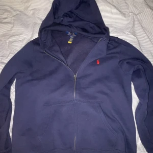 Mörkblå hoodie från Ralph Lauren - Säljer en snygg mörkblå hoodie från Ralph Lauren med dragkedja och huva. Den har en liten röd logga på bröstet och är perfekt för en avslappnad stil. Passar bra till både jeans och joggers. 🧢