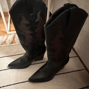 Svarta cowboy boots i skinn st 37 - Säljer ett par snygga svarta cowboy boots i skinn med spetsig tå och klack. De har coola broderade detaljer på skaftet som ger en unik look. Perfekta för höst och vinter, och funkar lika bra till vardags som till fest/festival. Skorna är i mycket bra skick och redo för nya äventyr! Storlek 37 köpta från Bershka i Polen, super sköna inuti!