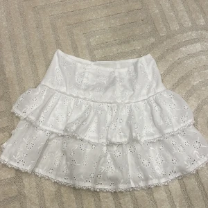 Vit kjol med volanger - Charmig vit kjol med dubbla volanger och broderade blommönster. Perfekt för en somrig look med sin luftiga design och romantiska stil. Passar bra till både enkla toppar och mer eleganta blusar.