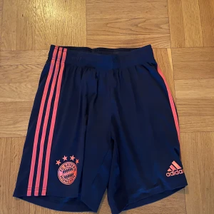 Blå fotbollsshorts från Adidas - Snygga blå fotbollsshorts från Adidas med rosa detaljer och Bayern München-logga. Perfekta för fotbollsträningen eller att bara chilla i. De har en elastisk midja för extra komfort.