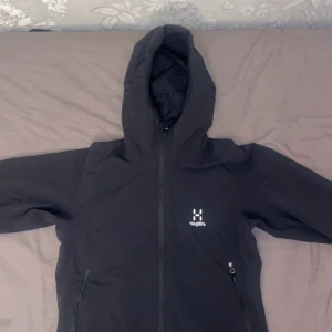 Svart vindjacka från Haglöfs - Säljer en svart vindjacka från Haglöfs i storlek M för dam. Jackan har en trasig dragkedja framtill och en justerbar huva. Perfekt för blåsiga dagar och utomhusaktiviteter.
