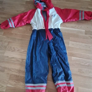 Regnoverall med huva - Säljer en regnoverall i rött, vitt och blått med huva och dragkedja. Regnoverallen har långa ärmar och elastisk midja för bättre passform. Perfekt för utomhusaktiviteter!