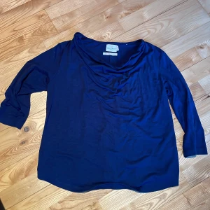 Mörkblå topp från Holly & Whyte - Säljer en stilren mörkblå topp från Holly & Whyte. Toppen har trekvartsärmar och en elegant draperad halsringning. Materialet är en blandning av polyester och viskos vilket ger en mjuk och bekväm känsla. Perfekt för en avslappnad men ändå chic look.