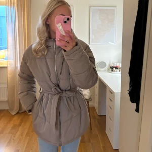 Beige pufferjacka med bälte - Säljer en snygg beige pufferjacka från Nelly. Jackan har en dragkedja framtill och ett bälte i midjan för en figursydd look. Perfekt för kyliga dagar med sin långa ärm och vadderade design.