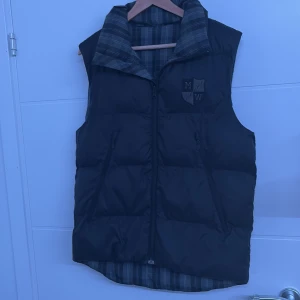 Svart dunväst från Marwin - Säljer en stilren svart dunväst från Marwin med ett broderat emblem på bröstet. Västen har en dragkedja framtill och ett rutigt foder som ger en snygg kontrast. Perfekt för kyliga dagar när du vill hålla värmen utan att kompromissa med stilen. Den kostade 1,588 från NK och den är ganska enkel att style och skön till våren och sommaren kan andvändas till hösten den är i storlek S men passar men jätte bra och kanske L beror på kroppsbygden, säljer på grund av flytt och rensar gaderoben 
