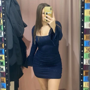 Mörkblå glittrig klänning - Snygg mörkblå klänning med glitterdetaljer och offshoulder-design. Klänningen har långa ärmar med volang och en tight passform som framhäver figuren. Perfekt för en kväll ute!