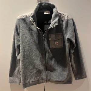 Grå fleecejacka från Moncler - Säljer en grå fleecejacka från Moncler med dragkedja och en liten bröstficka med logotyp. Jackan har långa ärmar och en hög krage. Perfekt för kyliga dagar och ger en stilren look.