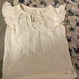 Vit blus från domitune - Säljer en söt vit blus från domitune med volangdetaljer på ärmarna och knytning vid halsen. Perfekt för en somrig look. Blusen har en liten metalllogga vid nederkanten.