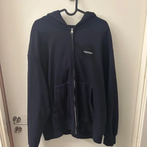 Mörkblå hoodie med dragkedja - Säljer en stilren hoodie med dragkedja och luva. Den har en liten vit logotyp på bröstet och praktiska fickor framtill. Perfekt för en avslappnad look.