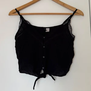 Svart spetsdetaljerad linnetopp - Svart linne från H&M med spetsdetaljer och knappar framtill. Toppen har smala axelband och en knytning vid nederkanten. 