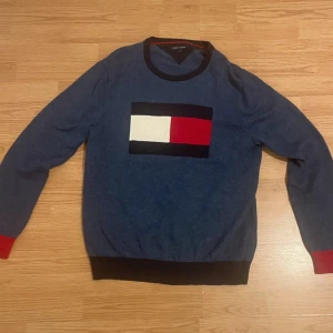 Blå tröja från Tommy Hilfiger - Säljer en snygg blå tröja från Tommy Hilfiger med deras ikoniska logotyp i rött, vitt och blått på bröstet. Tröjan har långa ärmar och ribbade muddar vid ärmslut och nederkant. Perfekt för en stilren look.