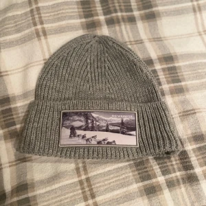 Grå stickad mössa med patch - Snygg grå stickad mössa från revenite med en cool patch framtill som visar ett vinterlandskap. Perfekt för kalla dagar och ger en stilren look. Passar både till vardags och utomhusaktiviteter. Aldrig använd.