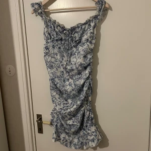  klänning  -  blommig klänning i vitt och blått med volangdetaljer. Klänningen har en smickrande passform och är perfekt för varma dagar. Den är ärmlös med breda axelband och har en lätt rynkad effekt vid bysten.