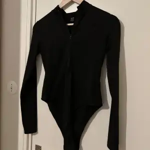 Snygg svart långärmad bodysuit med dragkedja framtill. Perfekt för en stilren look. Passar bra till både jeans och kjol. Helt oanvänd 