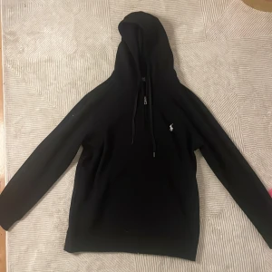 Svart hoodie från Ralph Lauren - Säljer en stilren svart hoodie från Ralph Lauren med klassisk logga på bröstet. Den har en justerbar huva med dragsko och långa ärmar. Perfekt för en avslappnad look. Storlek M/L men känns mest som M, har även en XXL vid intresse