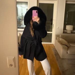 Pälskappa - Snygg svart kappa med stor huva och pälsdetaljer. Jackan har ett knytbälte i midjan för en elegant passform. Perfekt för kyliga dagar när du vill hålla stilen. Pälsen är avtagbar, storlek XS-S från femenina of sweden